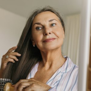 Beautiful, charming elderly female combing grey hair with round hairbrush standing against shelves in hallway or bedroom in front of mirror, looking at her reflection, getting ready to go to bed