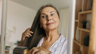 Beautiful, charming elderly female combing grey hair with round hairbrush standing against shelves in hallway or bedroom in front of mirror, looking at her reflection, getting ready to go to bed