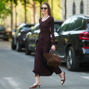 PARIS, FRANCE - APRIL 09: Alba Garavito Torre wears long, straight, brown hair with a middle part, natural complexion, subtle blush, and pink lipstick. Black rectangular sunglasses are worn. A brown tie-neck backless jersey long dress with long sleeves by Commense is styled with brown and white pointed slingback heels by Miu Miu. A mocha suede structured tote bag with double handles and gold clasp hardware by Demellier is carried by hand. A long leather necklace with a gold pendant and a delicate gold bracelet by Sempiterno Paris complete the look. Full body shot, during a street style fashion photo session, on April 09, 2025 in Paris, France. (Photo by Edward Berthelot/Getty Images)