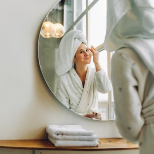 Caucasian woman with towel on head applies moisturizer for hyperpigmentation in front of mirror in bright white bathroom.