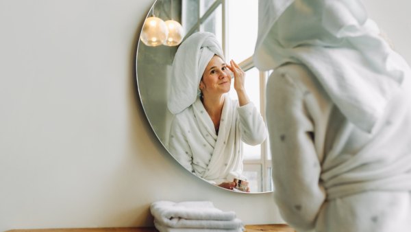 Caucasian woman with towel on head applies moisturizer for hyperpigmentation in front of mirror in bright white bathroom.