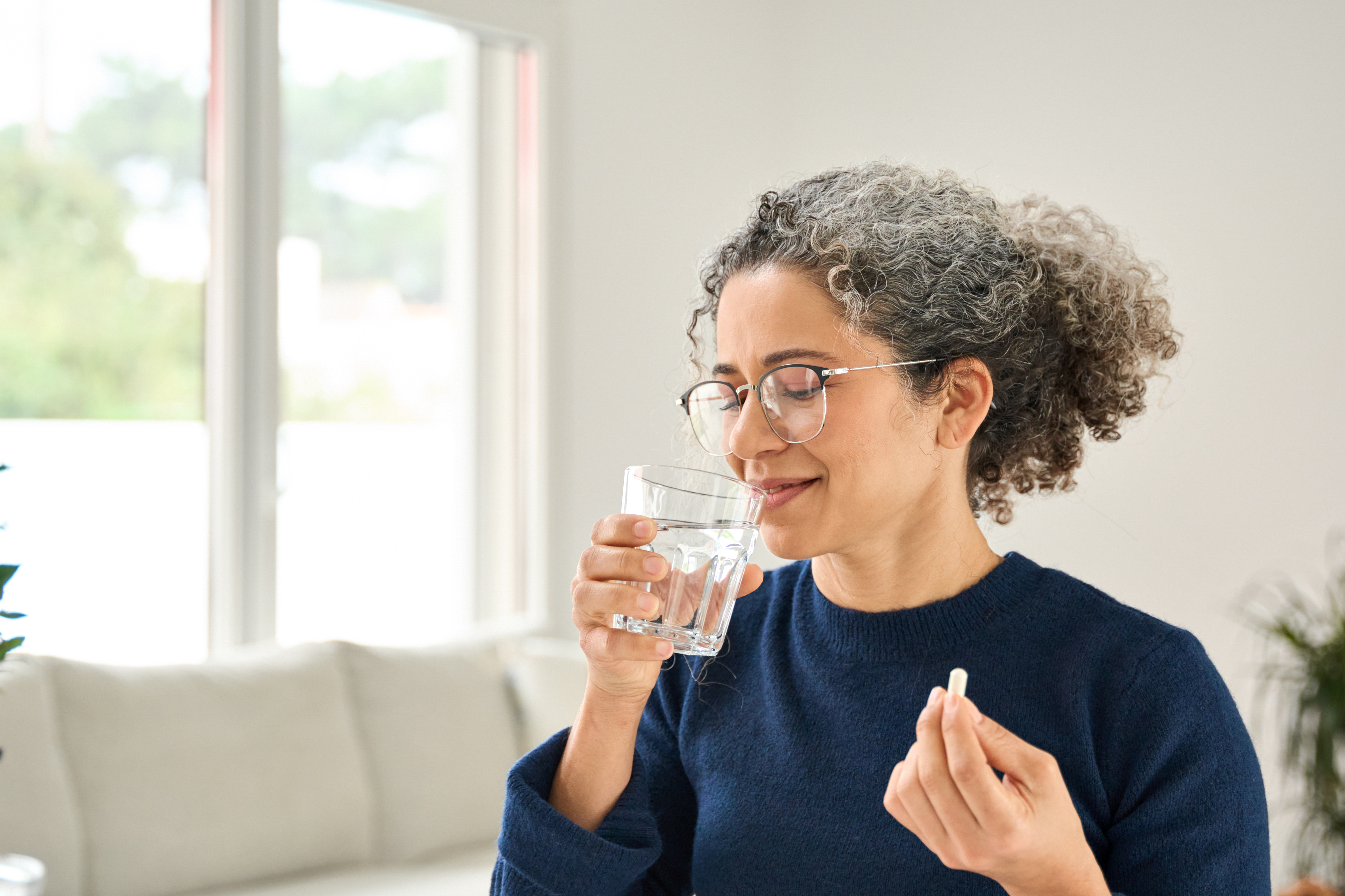 Happy healthy woman of middle age talking pill holding glass of water standing at home. Mature 50 years old lady taking vitamin health care supplement morning daily nutrition treatment concept.