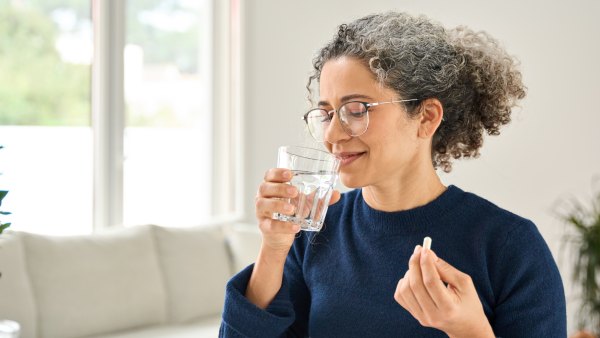 Happy healthy woman of middle age talking pill holding glass of water standing at home. Mature 50 years old lady taking vitamin health care supplement morning daily nutrition treatment concept.