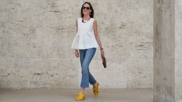 Alba Garavito Torre wears black sunglasses, long straight brown hair with a center part, a white pleated peplum sleeveless blouse by Commense, and blue high-rise raw-hem denim jeans. A yellow clog sandal with floral detailing by AGL is worn, paired with a jean denim and brown mini Jackie handbag by Gucci. Accessories include a chunky necklace with colorful natural stones by Sempiterno Paris and gold jewelry. Full body shot, during a street style fashion photo session, on May 02, 2025 in Paris, France.