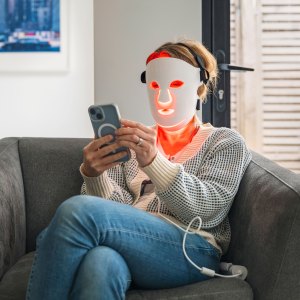 A mature woman uses her red LED face mask (red light therapy) whilst relaxing at home in an armchair. She uses her smartphone - stock photo