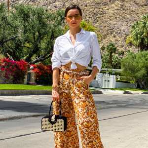 PARIS, FRANCE - APRIL 11: Julia Comil wears floral print flared flowy orange and brown palazzo pants by Jonathan Simkhai, a white linen shirt by Sezane, sustainable mules shoes in biodegradable nappa leather and natural wood by Nicholas Kirkwood, a vintage inspired cane box handbag by Mehry Mu, orange aviator sunglasses by ferragamo, during an online remote street style fashion photo session via Apple iphone / Facetime and the CLOS app as the model is based in Los Angeles - California and the photographer in Paris - France, on April 11, 2021 in Paris, France. (Photo by Edward Berthelot/Getty Images)