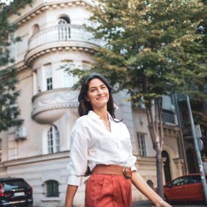 A confident woman strolls down a city street lined with lush trees and a historic building, exuding positivity and independence. The scene captures a blend of modern life and architectural charm.