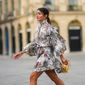 Alexandra Pereira wears gold and pearl earrings, a white with burgundy and pale colors print pattern belted turtleneck dress from Zimmerman with puffy sleeves and burgundy buttons, silver and diamonds rings, gold bracelets from Piaget, a gold shiny leather handbag / clutch from Bottega Veneta, on June 21, 2021 in Paris, France.
