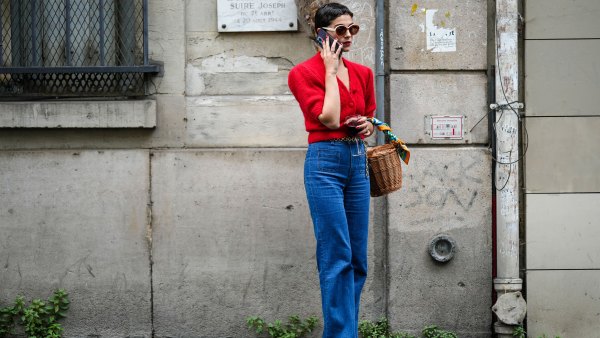 A guest wears circle sunglasses, a red fluffy puffy short sleeves / buttoned cropped cardigan, navy blue denim wide legs pants, a brown wickers basket handbag, a green / blue / orange print pattern silk scarf, rings, white shiny varnished leather heels shoes, a gold chain belt, outside the Hermes show, during Paris Fashion Week - Menswear Spring/Summer 2023, on June 25, 2022 in Paris, France.