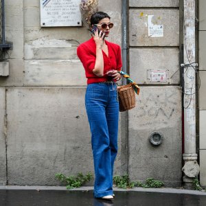 A guest wears circle sunglasses, a red fluffy puffy short sleeves / buttoned cropped cardigan, navy blue denim wide legs pants, a brown wickers basket handbag, a green / blue / orange print pattern silk scarf, rings, white shiny varnished leather heels shoes, a gold chain belt, outside the Hermes show, during Paris Fashion Week - Menswear Spring/Summer 2023, on June 25, 2022 in Paris, France.