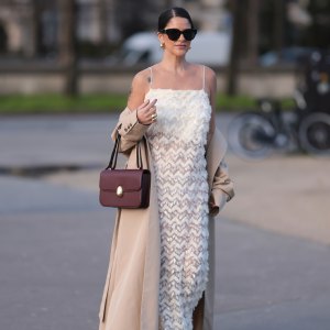 PARIS, FRANCE - FEBRUARY 27: Gili Biegun seen wearing YSL black sunglasses, Bottega Veneta gold tear drops earrings, white long sheer pearl embroidered maxi dress, gold statement rings, beige oversized long coat, Neous burgundy leather bag and burgundy varnished leather pumps / heels, outside Anrealage, during the Womenswear Fall/Winter 2024/2025 as part of Paris Fashion Week on February 27, 2024 in Paris, France. (Photo by Jeremy Moeller/Getty Images)