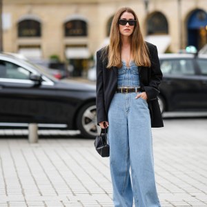 PARIS, FRANCE - SEPTEMBER 29: A guest wears black sunglasses, gold earrings, a blue denim embroidered seams heart-neck / corset top, a black oversized blazer jacket, a black shiny leather belt, blue denim large wide legs pants, black shiny leather handbag, black shiny leather ankle boots , outside Chloe, during Paris Fashion Week - Womenswear Spring/Summer 2023, on September 29, 2022 in Paris, France. (Photo by Edward Berthelot/Getty Images)
