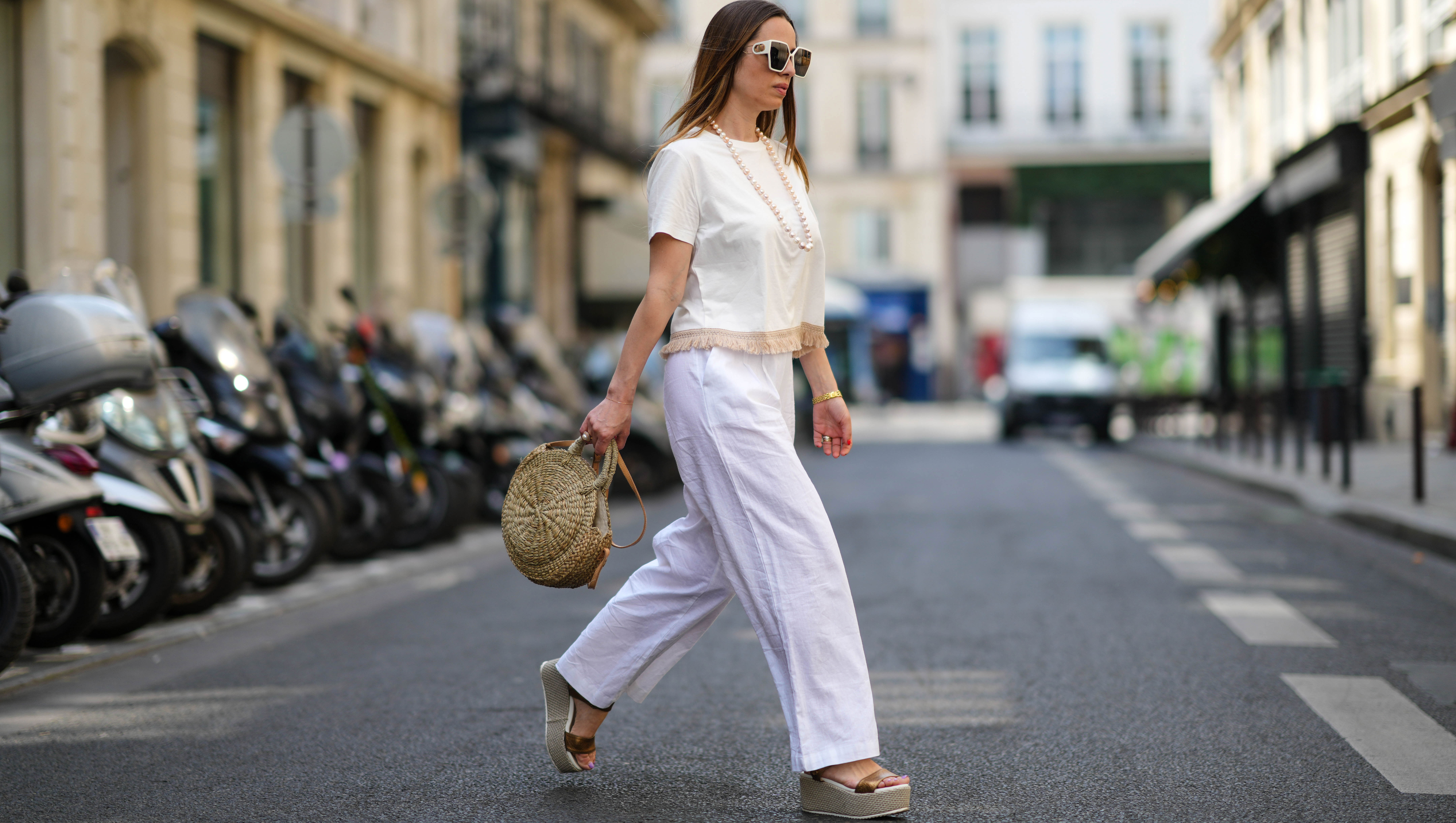 PARIS, FRANCE - MAY 10: Maria Rosaria Rizzo wears white sunglasses, a white pearls long necklace, a white latte t-shirt with fringed borders, a beige wickers handbag from Guerlain, a gold large bracelet, gold rings, high waist white wide legs linen pants, gold shiny leather strappy with wicker wedge heels sandals, during a street style fashion photo session, on May 10, 2022 in Paris, France. (Photo by Edward Berthelot/Getty Images)