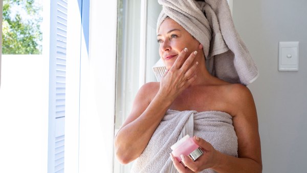 Smiling woman applying moisturizer on face standing by window at home - stock photo
