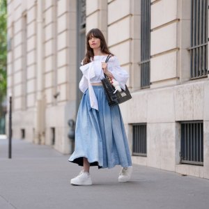 PARIS, FRANCE - JULY 18: Marion Félicité wears a white cropped top with oversized puffed sleeves, gathered seams and an asymmetrical bow detail on the chest by Patou, paired with a high-waisted, voluminous light blue denim maxi skirt with washed effect. A black and beige woven leather tote bag by Lancel is carried by hand, customized with a "Surprise Shake" Labubu Coca-Cola plush charm and colorful enamel keyrings. White Buffalo platform sneakers with lace-up closure are adorned with decorative silver and pearl charms including a butterfly and floral motif. Multicolored beaded bracelets are worn on the wrist. Fingernails are painted in a glossy gradient pink with green. Hair is styled in long, loose waves with front bangs and a side part. during a street style fashion photo session, on July 18, 2025 in Paris, France. (Photo by Edward Berthelot/Getty Images)