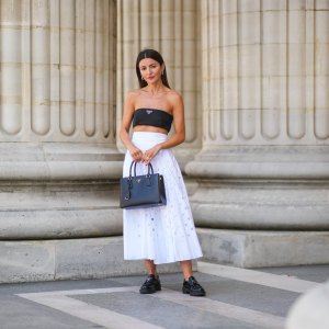 PARIS, FRANCE - JUNE 21: Alexandra Pereira wears silver and gold earrings, a black Prada shoulder-off crop-top, a white high accordion / gathered / pleated midi skirt with round holes from Prada, a black grained leather Galleria from Prada handbag, silver and diamonds rings, black shiny leather loafer shoes from Prada, on June 21, 2021 in Paris, France. (Photo by Edward Berthelot/Getty Images)