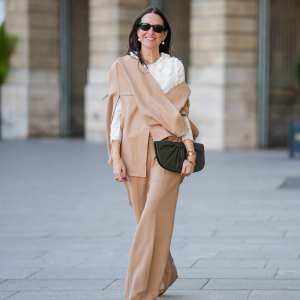 Alba Garavito Torre wears black sunglasses from Ray Ban, white large pearls earrings, a white shirt with cut-out lace pattern large collar, a beige felt / wool oversized cloak, matching beige wool / felt palazzo pants, a black shiny leather ruffled handbag, a gold Love bracelet from Cartier, gold bracelet, gold rings, brown leather pointed pumps heels shoes, during a street style fashion photo session, on October 28, 2021 in Paris, France