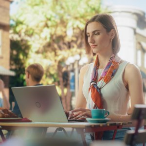 Female Freelancer Using Laptop Computer for Remote Online Work in a Cafe. Young Creative Woman Having Breakfast Croissant with Coffee while Teleworking From a Terrace on a Sunny Summer Day