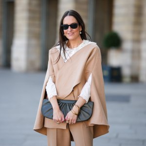 Alba Garavito Torre wears black sunglasses from Ray Ban, white large pearls earrings, a white shirt with cut-out lace pattern large collar, a beige felt / wool oversized cloak, matching beige wool / felt palazzo pants, a black shiny leather ruffled handbag, a gold Love bracelet from Cartier, gold bracelet, gold rings, during a street style fashion photo session, on October 28, 2021 in Paris, France.