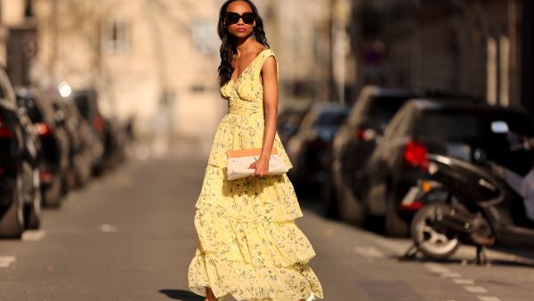 Emilie Joseph @in_fashionwetrust wears black sunglasses, a pale yellow with green and yellow flower print pattern V-neck / tank-top / backless / belted / ruffled maxi long dress, a beige fabric ruffled clutch, silver rings, pale yellow matte leather pointed pumps heels shoes, during a street style fashion photo session, on March 26, 2022 in Paris, France.