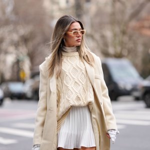 A guest wears yellow sunglasses, cream turtleneck long sleeve shirt, white pattern oversized buttoned up long sleeve shirt, cream knit oversized sweater, cream oversized wool coat, white mini skirt, outside Altuzarra, during the New York Fashion week Fall/Winter 2025 on February 8, 2025 in New York, New York.