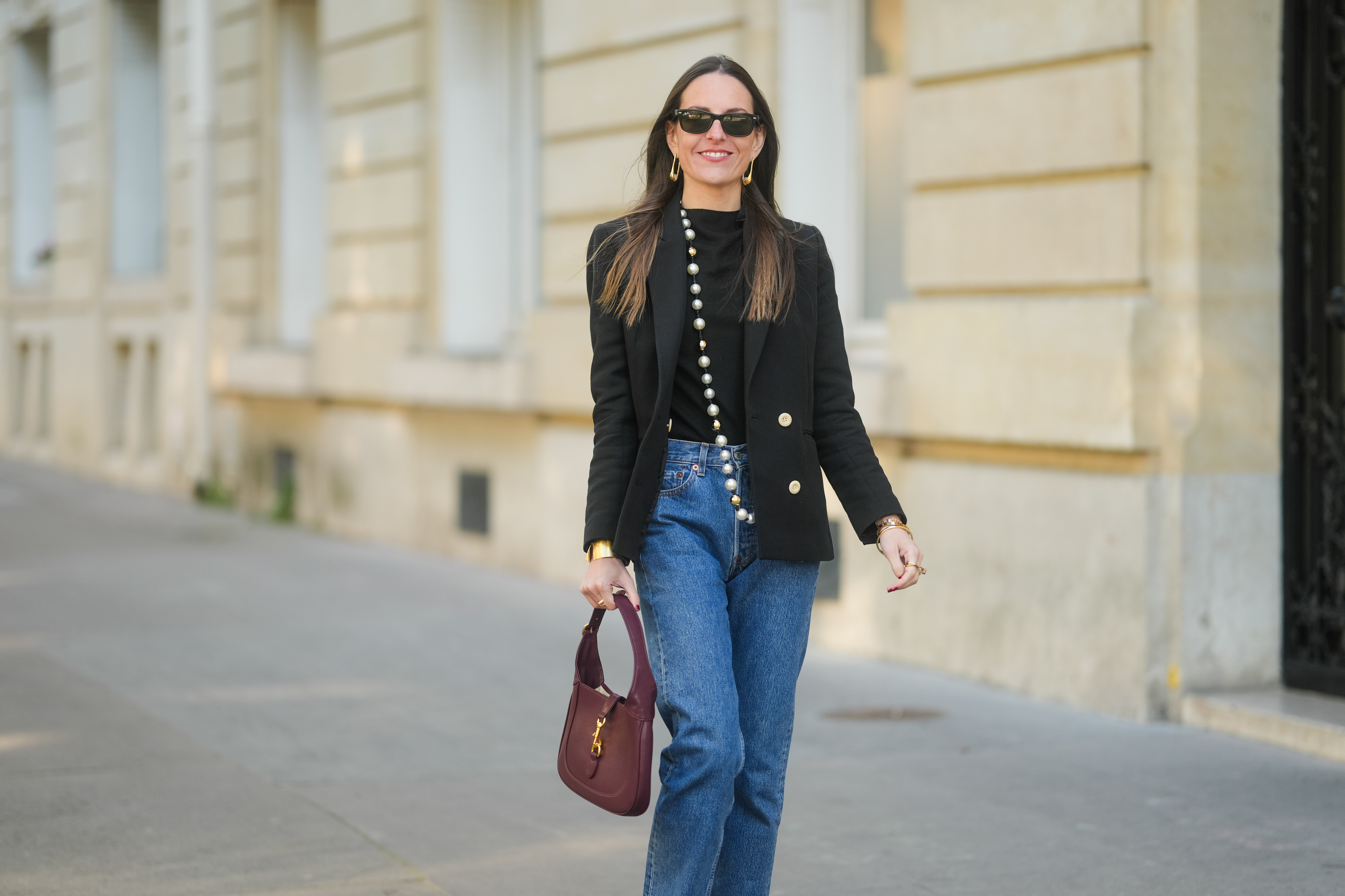 Alba Garavito Torre wears long straight brown hair styled with a center part. Black rectangular sunglasses are styled with large gold geometric earrings. A black T-shirt is layered under a tailored black double-breasted blazer with gold buttons by Sandro. A long strand of oversized pearls worn as a necklace by Sempiterno Paris is draped down the front. Classic blue Levi’s 5015 straight-leg jeans are worn with the hem grazing the top of the shoe. A burgundy leather Jackie bag by Gucci is carried. Full body shot, during a street style fashion photo session, on April 09, 2025 in Paris, France.