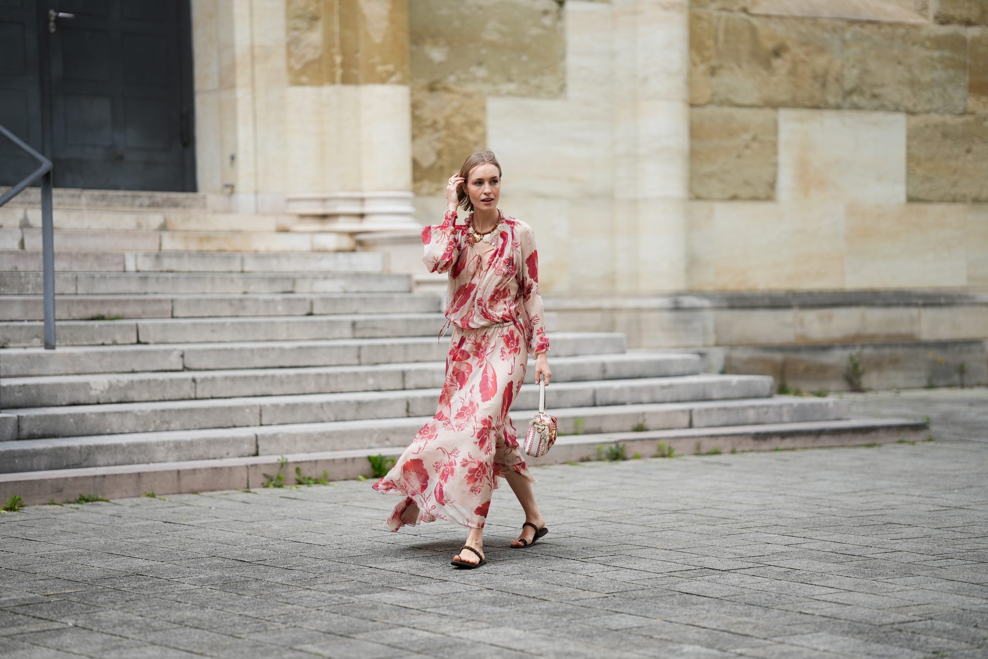 MUNICH, GERMANY - MAY 29: Marlies Pia Pfeifhofer is seen wearing a light beige silk chiffon long sleeve maxi dress with red and pink prints of feathers and flowers, drawstring waist, deep V-neck with front tie and smocking details on the collar and cuffs from FENDI, a natural-colored Baguette canvas bag with coral bandana embroidery, small fringes on the flap, calfskin leather straps, golden hardware and logo detail as well as an additional golden chain metal charm with marine-inspired pendants and a red, pink and white bandana charm with golden logo clip from FENDI, a pair of chocolate brown leather gladiator sandals with a braided leather toe strap and one main strap from Alex Rivière x Manebi, a gold-colored metal and brown nappa leather Forever Fendi necklace with shell-shaped pendants from FENDI, a yellow golden Leaves ring from Ole Lynggaard on May 29, 2025 in Munich, Germany. (Photo by Moritz Scholz/Getty Images)