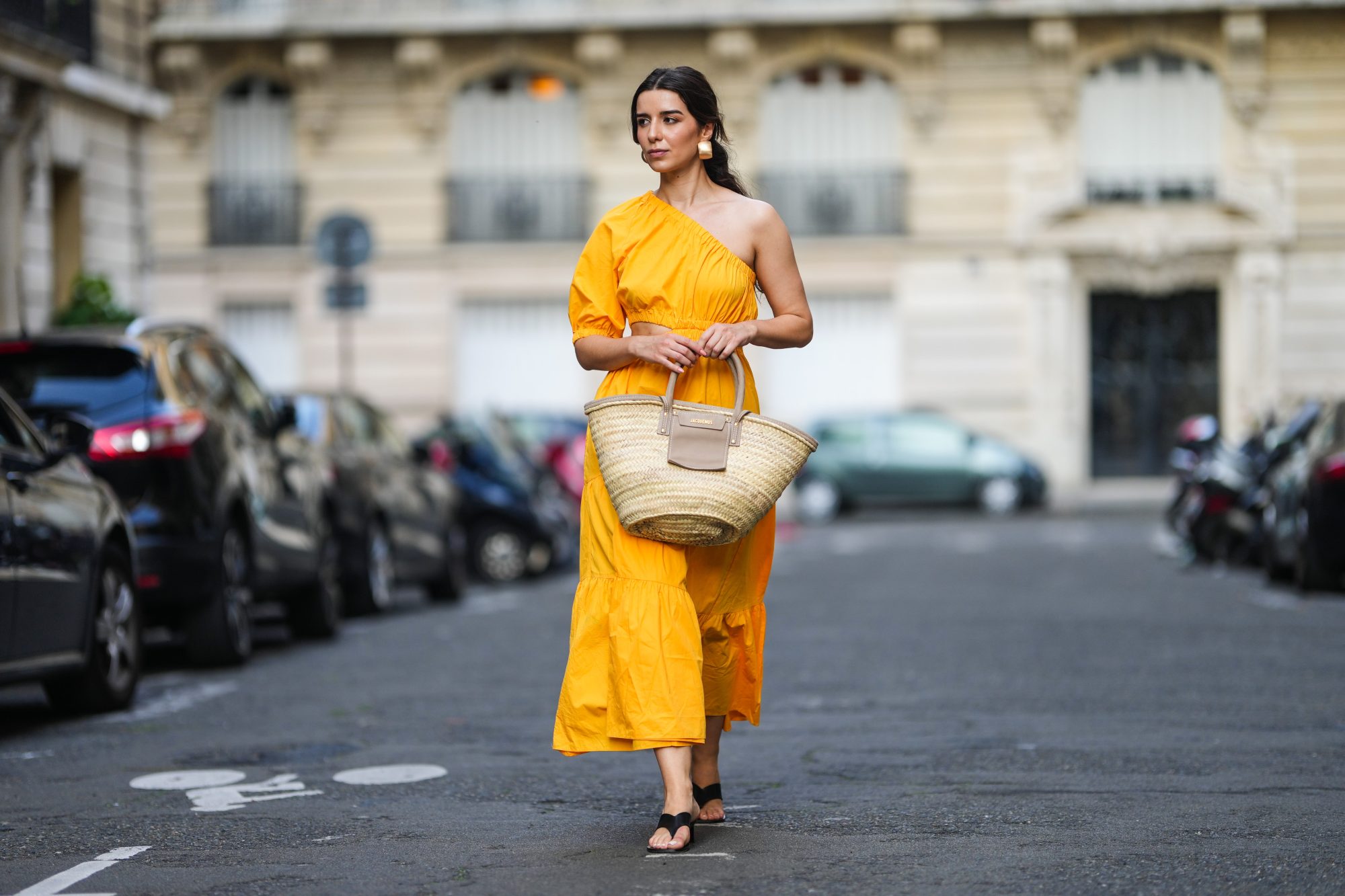 PARIS, FRANCE - AUGUST 12: Ketevan Giorgadze @katie.one wears a one shoulder cut out maxi dress in orange from Mango, oversized golden hoop earrings from Zara, flip flop heeled black sandals shoes from APC Paris, a Le Grand Panier beach straw bag by Jacquemus, on August 12, 2021 in Paris, France. (Photo by Edward Berthelot/Getty Images)