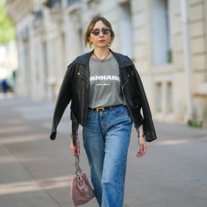PARIS, FRANCE - APRIL 09: Emy Venturini wears a black leather jacket with wide lapels and zipped pockets, layered over a faded grey graphic T-shirt by Zara featuring “WANNABE” and the Spice Girls logo. Blue high-waisted jeans by The Frankie Shop and a black belt with a gold H buckle by Hermes are visible. Gold sculptural earrings by Rabanne and a red beaded necklace complete the look. Mid shot, during a street style fashion photo session, on April 09, 2025 in Paris, France. (Photo by Edward Berthelot/Getty Images)