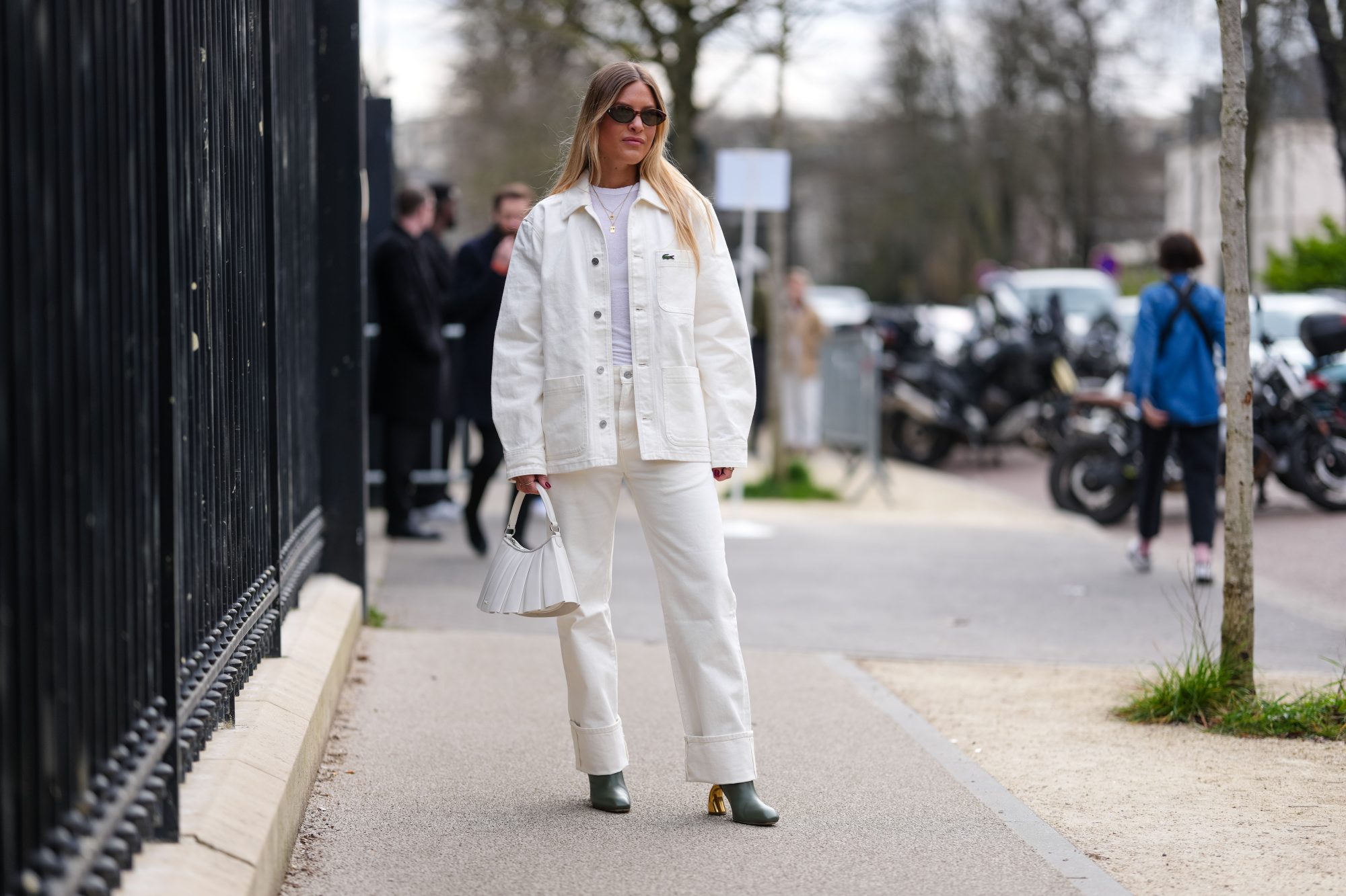 PARIS, FRANCE - MARCH 09: A guest wears dark brown sunglasses, white t-shirt, gold layered necklaces, cream oversized denim jean Lacoste jacket, cream loose denim jean cuffed Lacoste pants, shiny white Lacoste leather bag, dark green heeled leather shoes, outside Lacoste, during the Paris Fashion week Women's Fall/Winter 2025-2026 on March 9, 2025 in Paris, France. (Photo by Edward Berthelot/Getty Images)