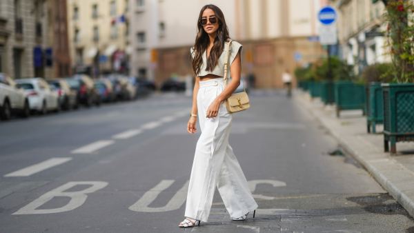 Tamara Kalinic wears gold Bottega Veneta sunglasses, a gold chain pendant necklace, a pale gray Jacquemus V-neck collar crop top with epaulets, white large flared Philosophy belted trousers pants, a gold Juste Un Clou Cartier bracelet, a gold Love Cartier bracelet, gold and silver Bee My Love Chaumet bracelets, a gold ring, a beige shiny leather Saint Laurent YSL shoulder bag, white shiny leather strappy heels Balenciaga sandals shoes, on April 29, 2021 in Paris, France.