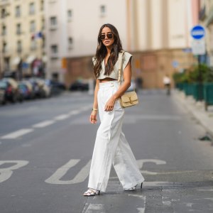 Tamara Kalinic wears gold Bottega Veneta sunglasses, a gold chain pendant necklace, a pale gray Jacquemus V-neck collar crop top with epaulets, white large flared Philosophy belted trousers pants, a gold Juste Un Clou Cartier bracelet, a gold Love Cartier bracelet, gold and silver Bee My Love Chaumet bracelets, a gold ring, a beige shiny leather Saint Laurent YSL shoulder bag, white shiny leather strappy heels Balenciaga sandals shoes, on April 29, 2021 in Paris, France.