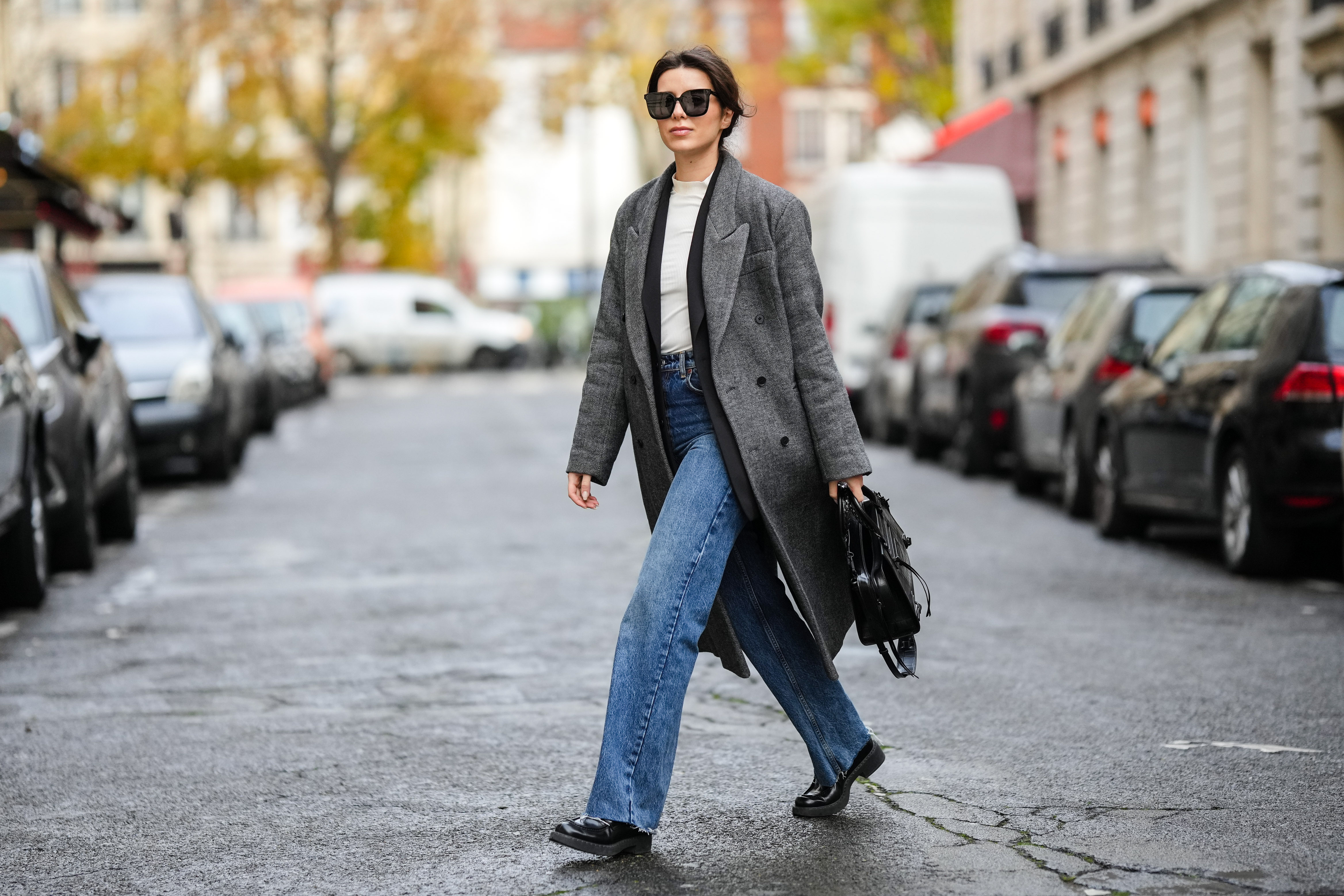 Katie Giorgadze @katie.one wears black sunglasses, a white latte ribbed high neck wool pullover, a black long blazer jacket, a black and white print pattern long oversized wool coat, high waist blue denim large ripped pants, a black shiny leather crocodile print pattern shoulder bag from Balenciaga, black shiny leather block heels loafers from Prada, on November 26, 2021 in Paris, France.