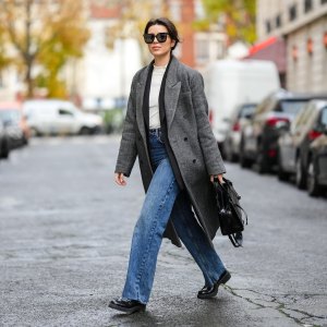 Katie Giorgadze @katie.one wears black sunglasses, a white latte ribbed high neck wool pullover, a black long blazer jacket, a black and white print pattern long oversized wool coat, high waist blue denim large ripped pants, a black shiny leather crocodile print pattern shoulder bag from Balenciaga, black shiny leather block heels loafers from Prada, on November 26, 2021 in Paris, France.