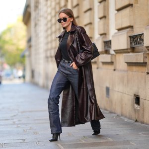 Diane Batoukina wears black sunglasses, a black t-shirt, a dark brown shiny leather long coat, a black shiny grained leather Kelly small belt from Hermes, black denim large pants, a black shiny leather Timeless handbag from Chanel, black shiny leather heels ankle boots , during a street style fashion photo session, on October 26, 2022 in Paris, France.