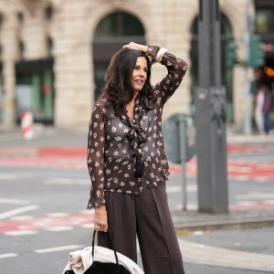 Angelika Hanel is seen wearing a chocolate brown flowing shirt with scarf collar, semi-sheer fabric, cream white floral print and button-up front from Massimo Dutti; a soft beige v-neck cashmere pullover with a relaxed silhouette and subtle ribbed cuffs and hem from Sa Su Phi; dark brown wide-leg linen blend trousers with a relaxed fit from H&M; a large black suede top-handle Soft Margaux 17 bag with softly structured sides, gold-tone buckles on the sides and a curved silhouette from The Row; a long black ceramic tassel cord necklace from Massimo Dutti; a golden necklace with a vintage Alhambra pendant in the shape of a cloverleaf from Van Cleef & Arpels; a golden Day-Date wristwatch from Rolex; a white gold pavé diamond tennis bracelet; an ecru colored Luca arm cuff set of two, made of synthetic resin from Sézane; a ring stacked with gold bands and a diamond from Marré; a diamond ring on July 27, 2025 in Frankfurt am Main, Germany.