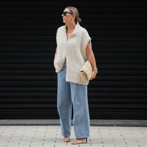 HAMBURG, GERMANY - SEPTEMBER 19: Aylin König is seen wearing a loose, chunky knit sleeveless, zip-up sweater in a soft beige tone with a wide collar and relaxed drape; light blue straight-leg jeans with a slightly wide fit; nude strappy heeled sandals; a beige woven-style clutch bag with a padded texture; stacked gold bracelets from Cartier; a watch with a metallic wristband and white clock face from Cartier; gold detailed rings; gold link earrings from Tiffany & Co HardWear Collection; black rectangular sunglasses; her dark hair is pulled back into a neat, low bun on September 19, 2025 in Hamburg, Germany. (Photo by Moritz Scholz/Getty Images)