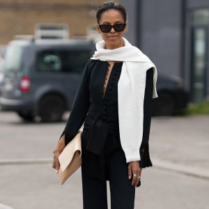 COPENHAGEN, DENMARK - AUGUST 7: Suzannà Gembege is seen wearing a white knit sweater draped over the shoulders; a black blouse with a deep V-neck big button details and fringe-accented sleeves; a matching black fringe-hem trousers; black flip flops; a black cat-eye sunglasses; a beige oversized clutch; silver rings; her black hair is styled straight in a ponytail outside the Marimekko show during Copenhagen Fashion Week day four on August 7, 2025 in Copenhagen, Denmark. (Photo by Moritz Scholz/Getty Images)