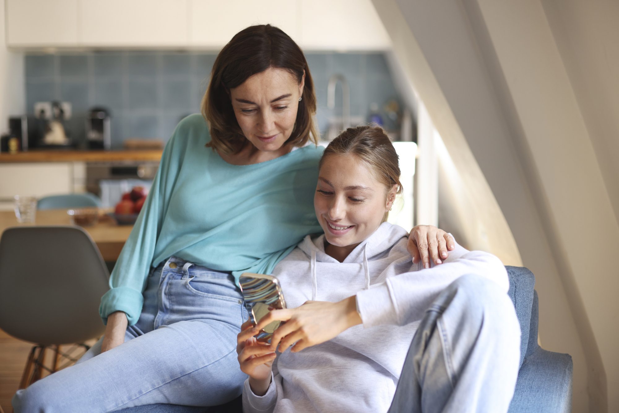 A mom and daughter spending time together, looking at a phone, in the living room. Photo by Catherine Delahaye/Getty Images