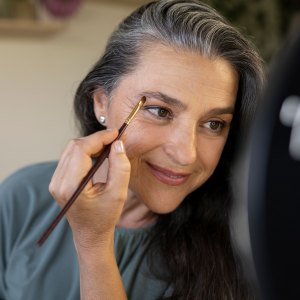 Smiling female vlogger brushing eyebrows during tutorial at home - stock photo