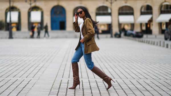 Emilie Joseph wears sunglasses, a khaki military jacket from Saint-Laurent YSL, a white shirt / blouse from Zara with ruffled collar, blue denim jeans pants from Levi's, brown high heeled knee-high pointy leather crocodile pattern boots from Paris Texas, on January 26, 2021 in Paris, France.