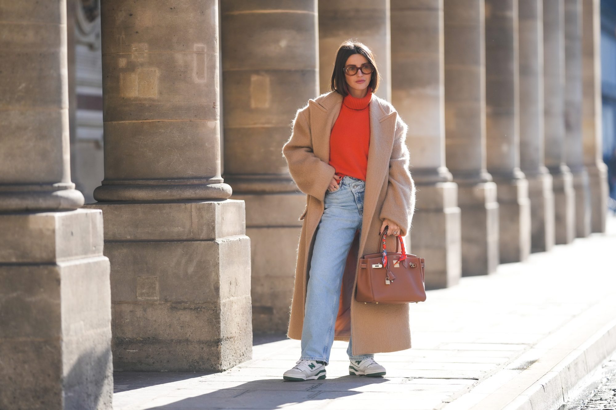 PARIS, FRANCE - FEBRUARY 09: Alexandra Pereira wears brown sunglasses, gold earrings, a neon orange turtleneck wool pullover, a beige oversized long winter coat, a brown shiny leather Birkin handbag from Hermes, diamonds ring, an orange / blue / yellow / white print pattern silk scarf from Hermes, blue faded denim large mom jeans pants, white and green leather sneakers from New Balance, during a street style fashion photo session, on February 09, 2022 in Paris, France. (Photo by Edward Berthelot/Getty Images)