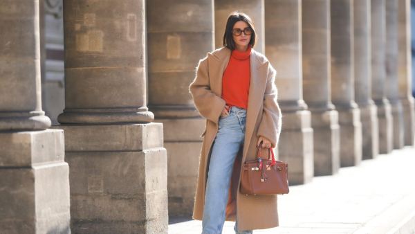 PARIS, FRANCE - FEBRUARY 09: Alexandra Pereira wears brown sunglasses, gold earrings, a neon orange turtleneck wool pullover, a beige oversized long winter coat, a brown shiny leather Birkin handbag from Hermes, diamonds ring, an orange / blue / yellow / white print pattern silk scarf from Hermes, blue faded denim large mom jeans pants, white and green leather sneakers from New Balance, during a street style fashion photo session, on February 09, 2022 in Paris, France. (Photo by Edward Berthelot/Getty Images)