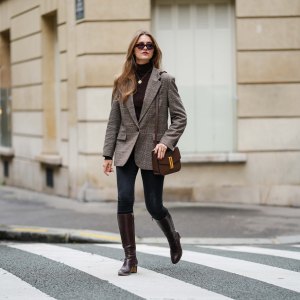 Segolene Hyppolite wears brown Jimmy Fairly sunglasses, gold necklace, grey checked Maje blazer jacket, a dark brown Maje turtleneck pullover, black denim jeans / pants, dark brown leather Rivecour boots, a brown leather Demellier London bag, during a street style fashion photo session, on October 12, 2024 in Paris, France.