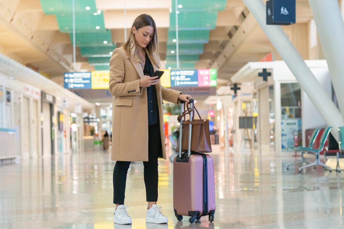 Woman looking at her smartphone to find her flight information while navigating an airport terminal with her luggage in tow.