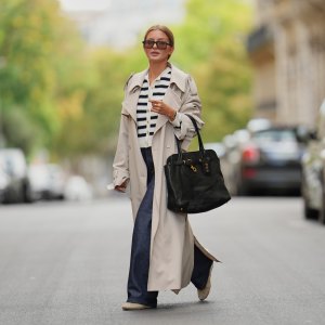 Diane Batoukina wears brown hair styled in a sleek low bun, oversized dark sunglasses by Kaleos, a beige trench coat by The Frankie Shop, a white and navy striped buttoned cardigan by APC, dark blue wide-leg jeans by APC, a black leather Miu Miu handbag with gold hardware, and beige and white sneakers by Miu Miu, during a street style fashion photo session, on September 05, 2025 in Paris, France.