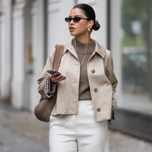 BERLIN, GERMANY - OCTOBER 08: Bella Emar with Iphone, Apple cable headphones wears Sézane Trench jacket, jumper knit & white light flared pants, flattered Boots, Liebeskind brown suede bag on October 08, 2025 in Berlin, Germany. (Photo by Christian Vierig/Getty Images)