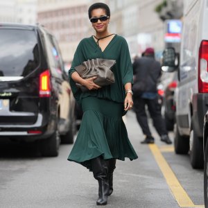 MILAN, ITALY - SEPTEMBER 27: Tamu McPherson wears a short natural black afro hairstyle, black rectangular sunglasses, small gold hoop earrings and a silver metal watch and bracelet, a taupe ruched leather pouch bag with gold-tone hardware handheld, a dark green flowing V-neck blouse with a tie at the neckline and dolman sleeves, a matching dark green gathered midi skirt with a tiered hem, black leather knee-high boots shoes with a block heel, outside Ferragamo, during Milan Fashion Week - Womenswear Spring/Summer 2026, on September 27, 2025 in Milan, Italy (Photo by Edward Berthelot/Getty Images)