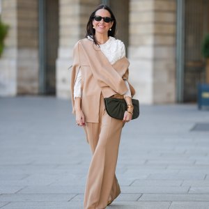Alba Garavito Torre wears black sunglasses from Ray Ban, white large pearls earrings, a white shirt with cut-out lace pattern large collar, a beige felt / wool oversized cloak, matching beige wool / felt palazzo pants, a black shiny leather ruffled handbag, a gold Love bracelet from Cartier, gold bracelet, gold rings, brown leather pointed pumps heels shoes, during a street style fashion photo session, on October 28, 2021 in Paris, France.