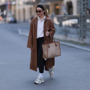 BERLIN, GERMANY - APRIL 09: Sonia Lyson seen wearing black cotton headband, Corlin Eyewear slim oval-shaped dark brown sunglasses, Worst Behavior white cotton polo sweater giving the look a sporty, laid-back vibe, Herskind long, cognac brown leather trench coat, Lululemon simple black comfortable and athleisure-inspired leggings, Gianni Chiarini large beige tote bag with brown leather trim and monogrammed initials, Nike white cotton crew socks and New Balance chunky cream white sneakers, on April 09, 2025 in Berlin, Germany. (Photo by Jeremy Moeller/Getty Images)